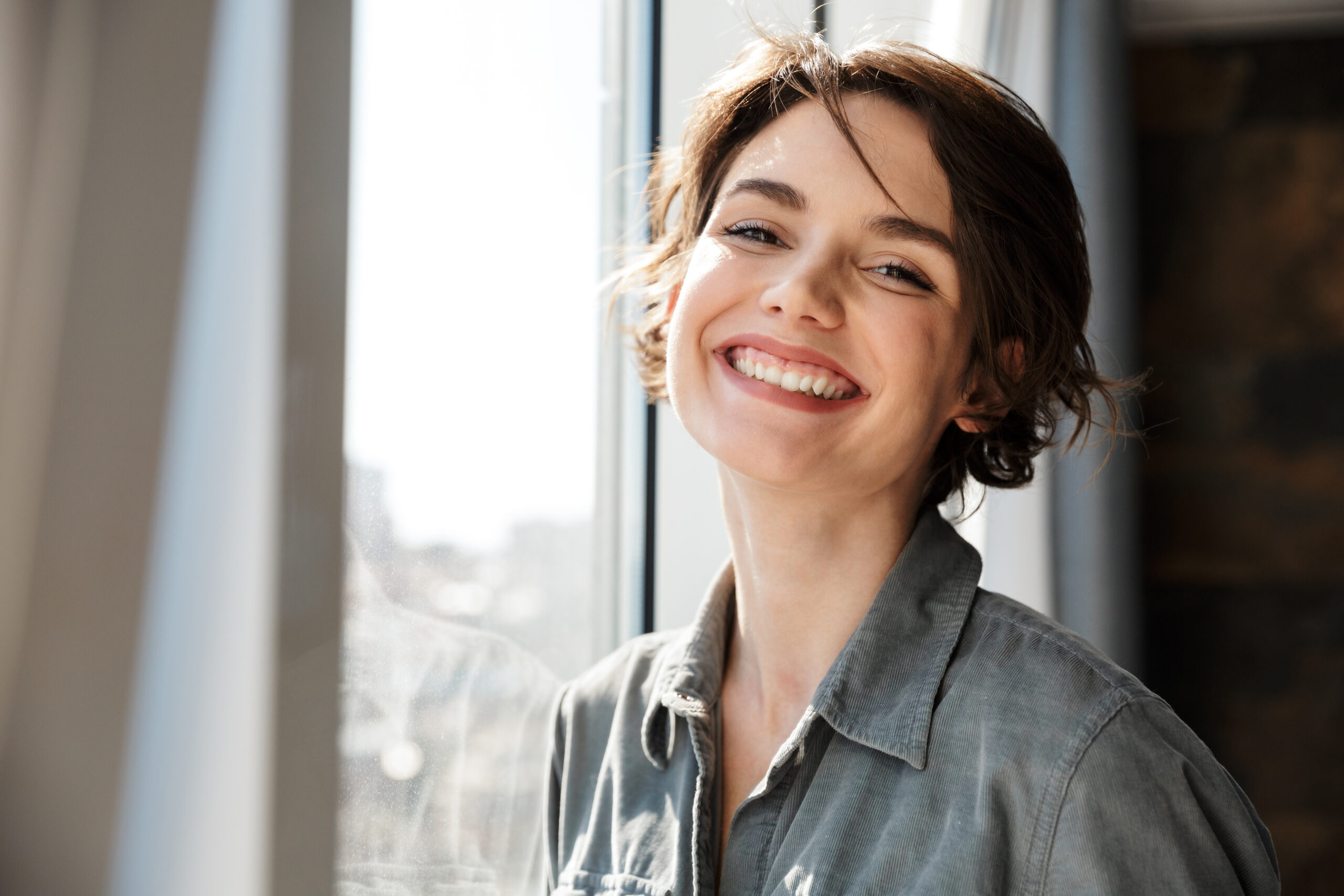 Image of beautiful young joyful woman smiling and looking at camera while standing near window